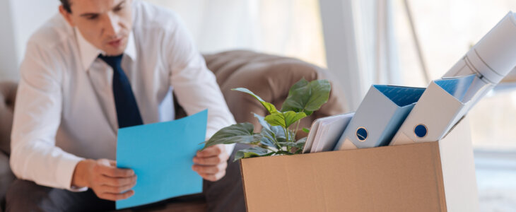 Important information. Concentrated calm young man sitting in an armchair and reading an important document after losing his job while a big box of personal things standing in front of him